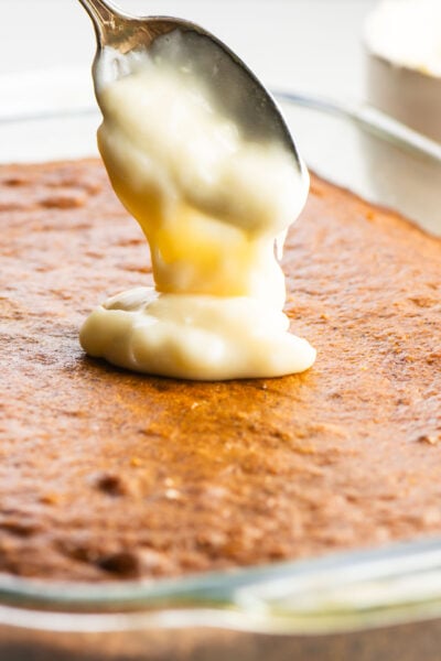A spoon pouring creamy sauce onto a baked dessert in a glass dish.