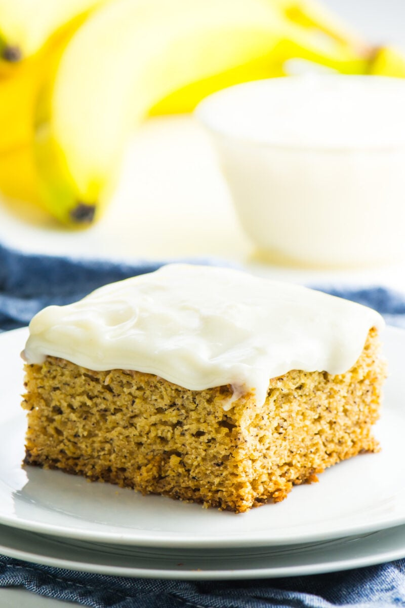 A slice of frosted vegan banana cake on a plate. In the background is more frosting and bananas.