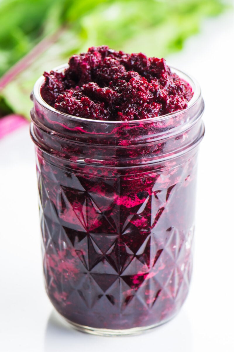 A jar holds pureed beets with beet greens behind it, with bright pink stems.