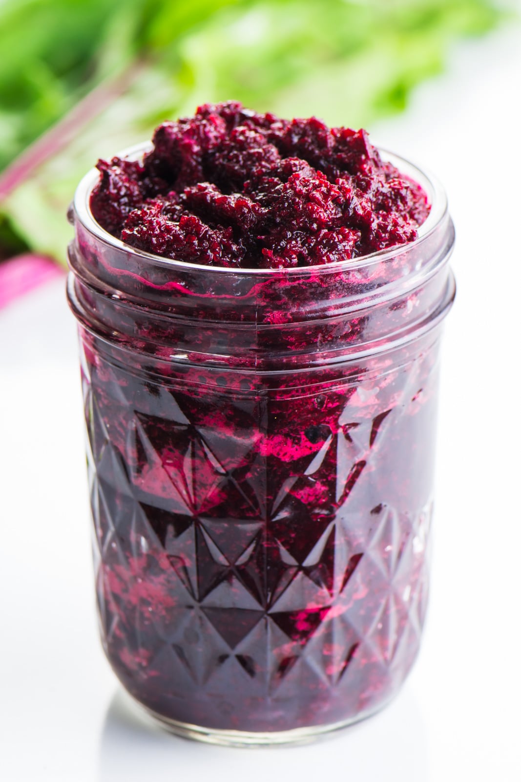 A jar holds pureed beets with beet greens behind it, with bright pink stems.