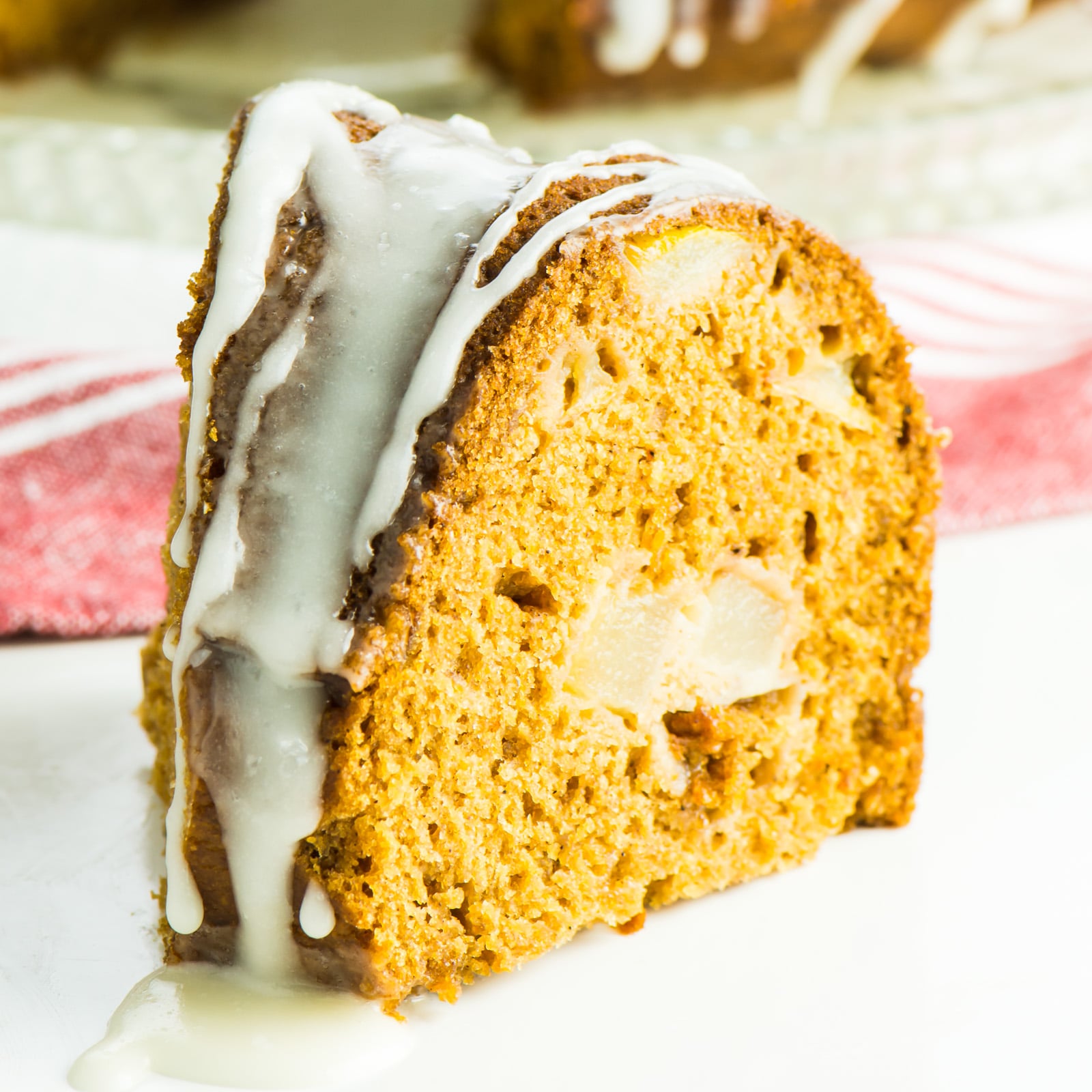 A slice of fresh pear cake sits on a white surface in front of a red and white kitchen towel.
