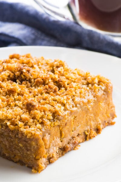 A vegan pumpkin pie bar sits on a white plate with a cup of coffee behind it.