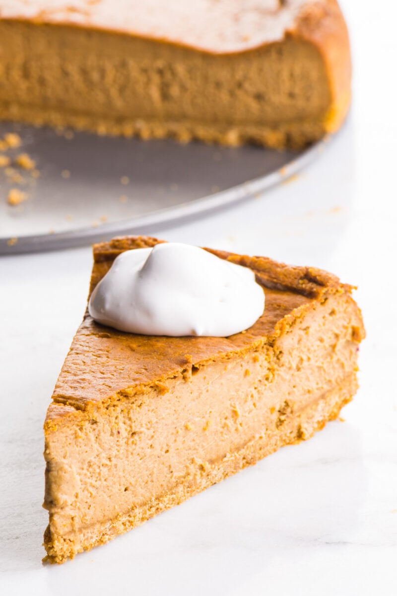 A slice of Vegan Pumpkin Cheesecake is served with vegan whipped cream on top. The rest of the cheesecake is behind it.