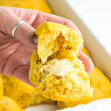 A hand holds a buttery bite of sweet potato dinner rolls with the pan behind it.