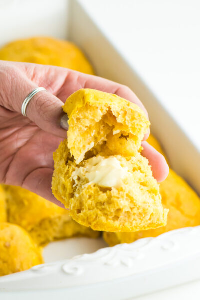 A hand holds a buttery bite of sweet potato dinner rolls with the pan behind it.