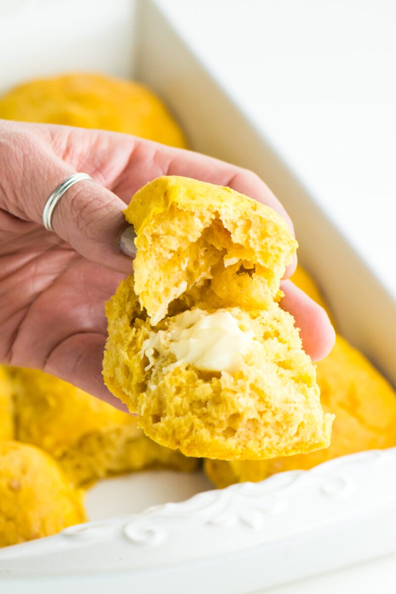A hand holds a buttery bite of sweet potato dinner rolls with the pan behind it.