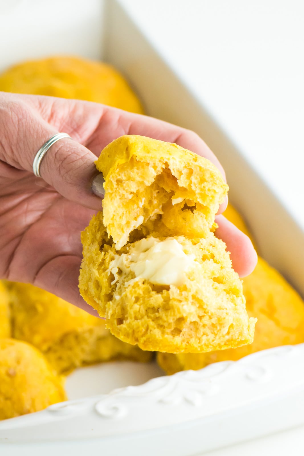 A hand holds a buttery bite of sweet potato dinner rolls with the pan behind it.