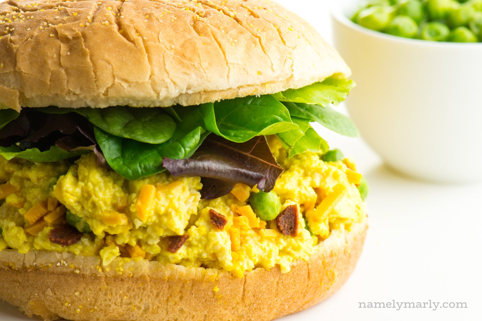 A bun has vegan egg salad along with greens. A bowl of frozen peas is in the background.