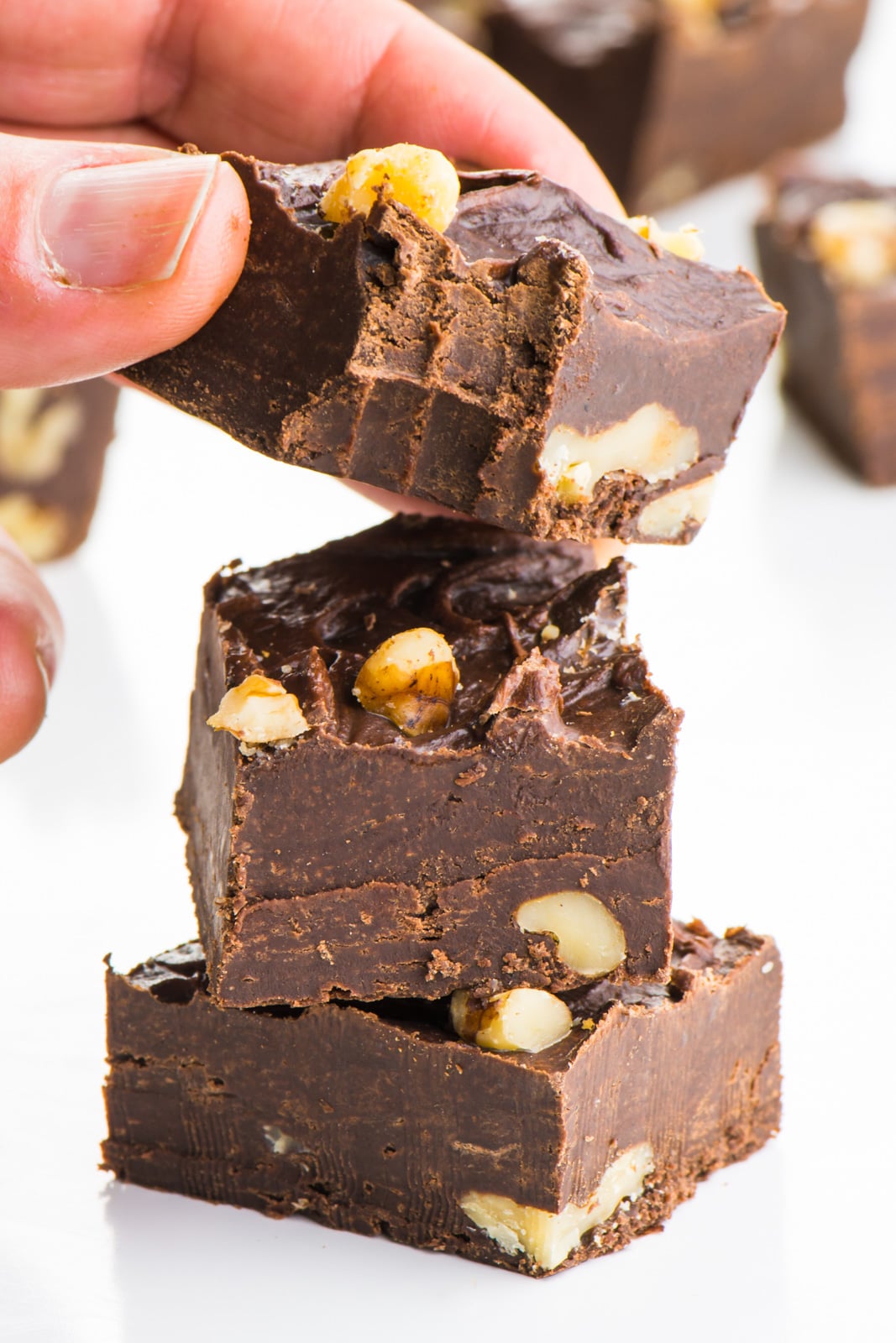 A hand holds a piece of chocolate fudge with a bite taken out, over a stack of more chocolate.