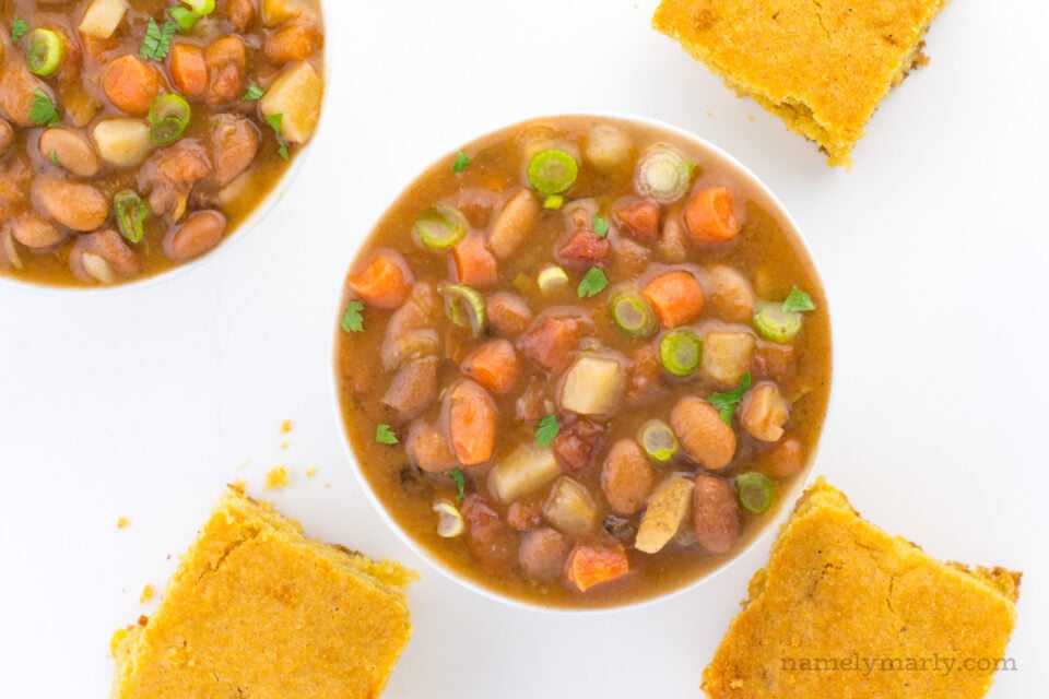 Two bowls of pinto bean soup are shown on a table, one is in the center and the other is only partially visible in the top left hand corner. There re slices of cornbread sitting around the bowls.