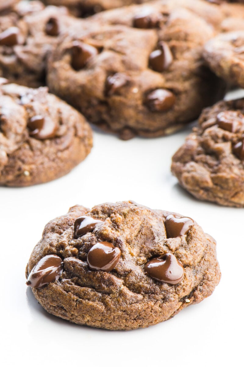 A single chocolate chocolate chip cookie sits in the foreground. There are several more cookies behind it.