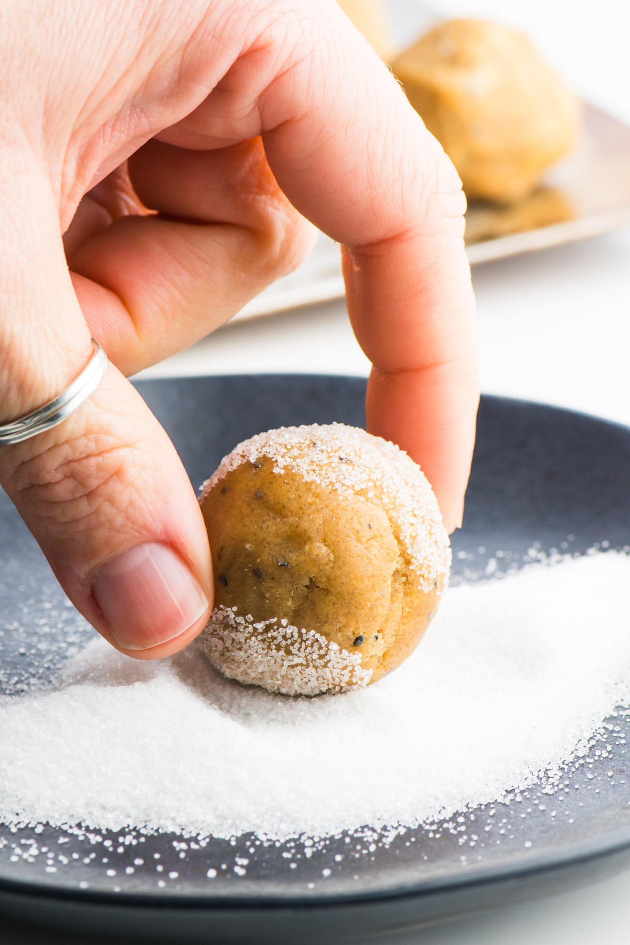 A hand is holding a vegan peanut butter cookie dough ball over a plate of sugar. The cookie dough ball has been partially rolled in the sugar.