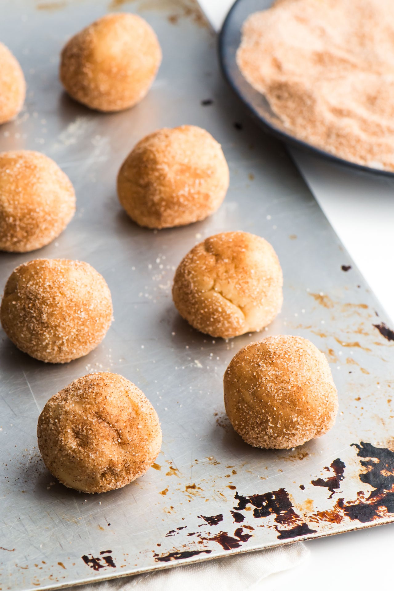Snickerdoodle cookie dough balls are lined up a cookie baking sheet, waiting to to in the oven. A plate of cinnamon sugar sits nearby.