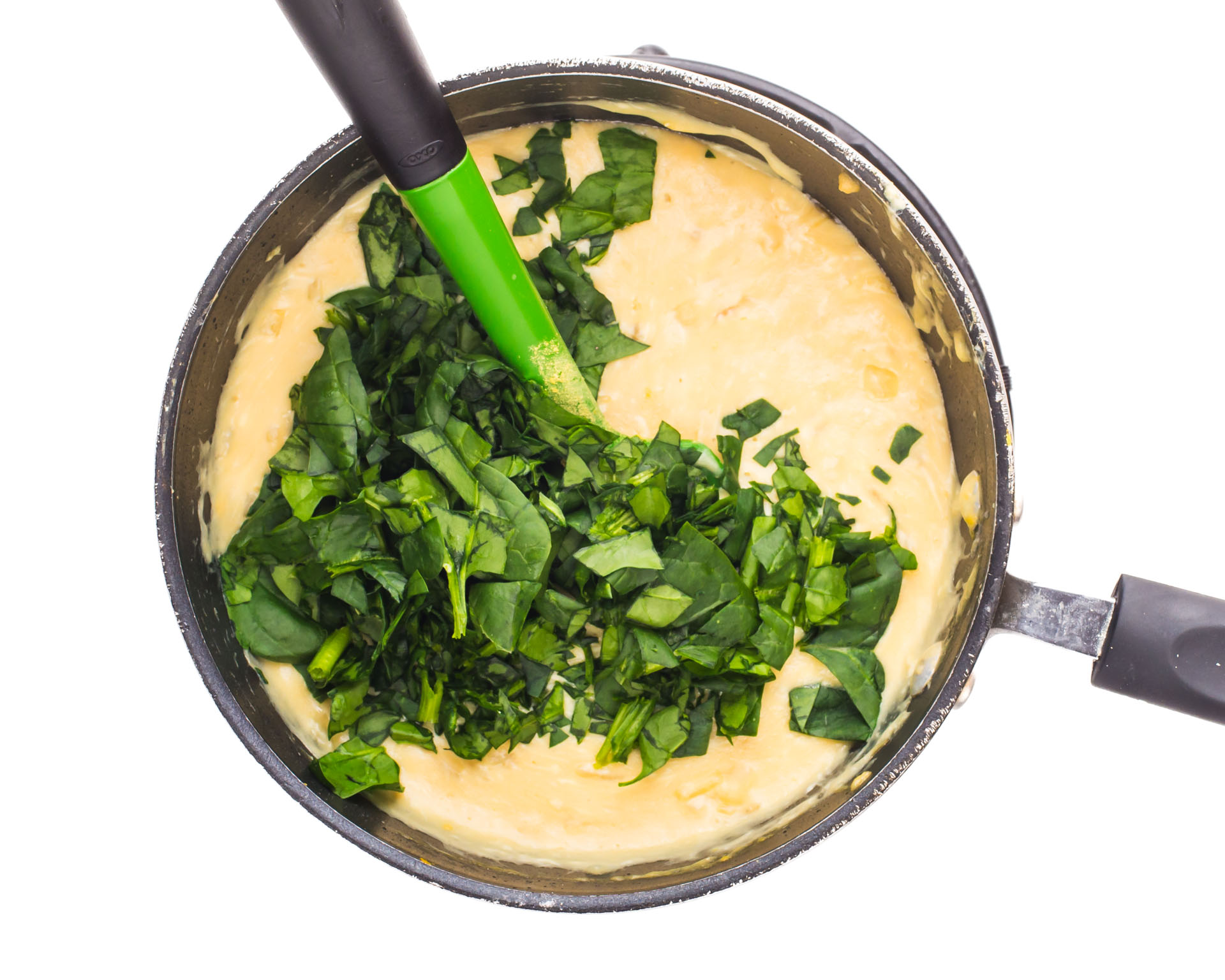 A saucepan shows the melted vegan cheese sauce with fresh spinach just added. A green spatula is in th epot, waiting to stir it all together.
