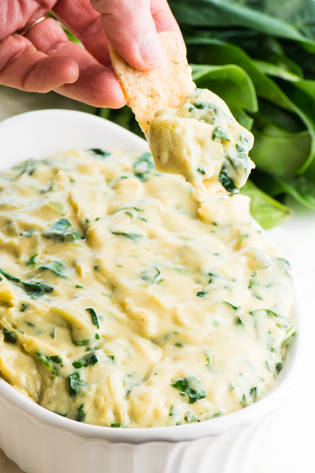 A hand holds a tortilla chip with vegan spinach artichoke dip with the bowl of dip behind it.