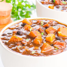 Two bowls of sweet potato chili sit one in front of the other. There are slices of sweet potatoes and a sprig of parsley in the background.