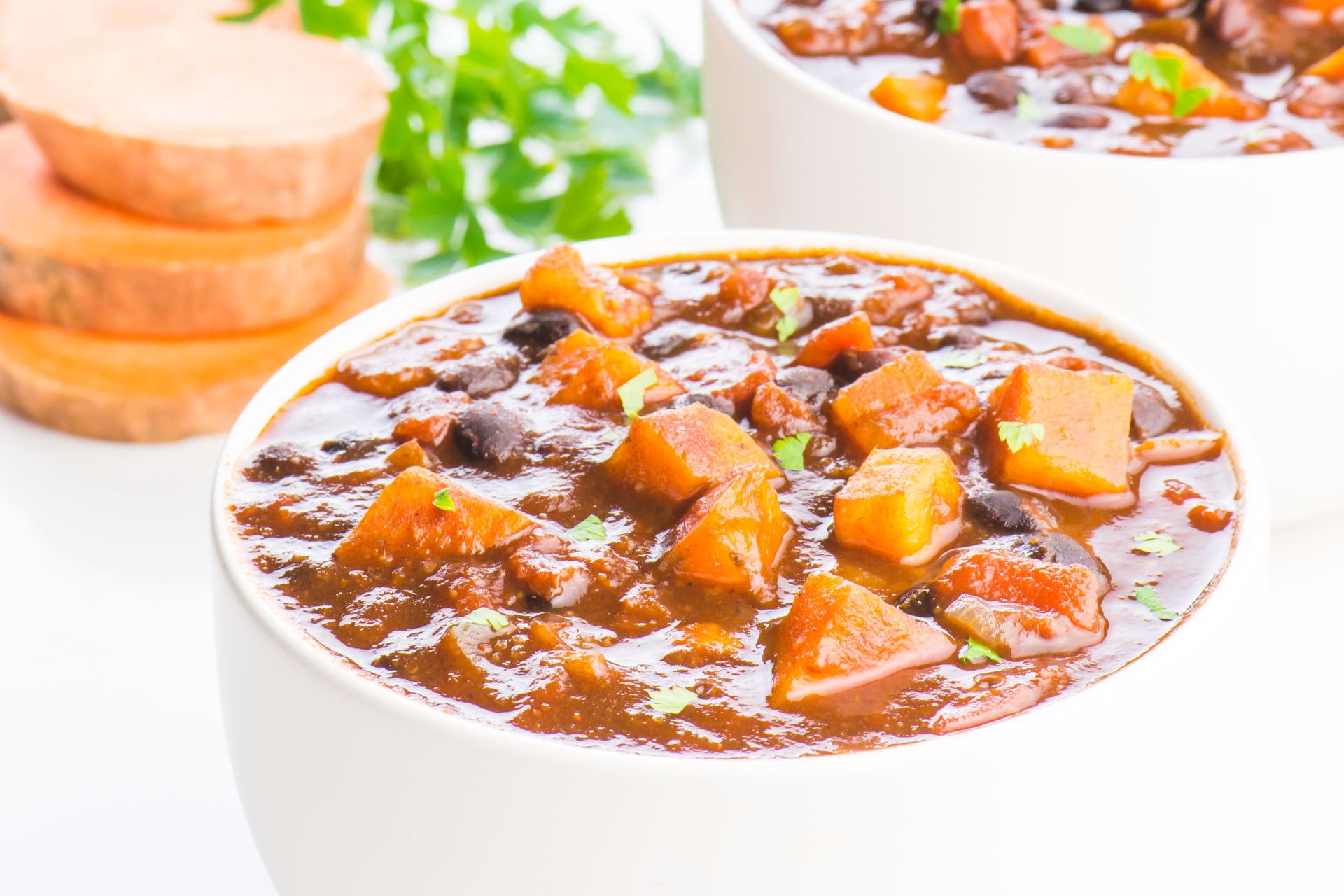 Two bowls of sweet potato chili sit one in front of the other. There are slices of sweet potatoes and a sprig of parsley in the background.