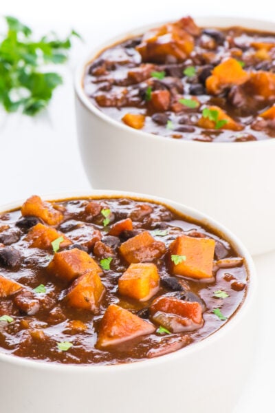 Two bowls of vegan sweet potato chili sit one in front of the other. A bit of parsley is in the background.