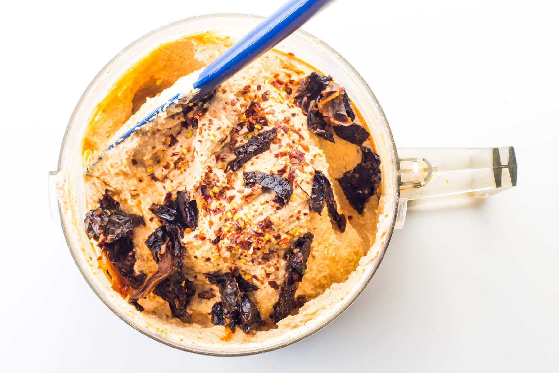 Creamy tofu is in a food processor, with sun-dried tomatoes, and spices on the top. There is a blue spatula on the side.