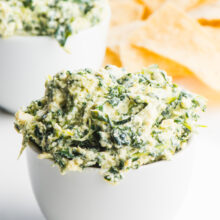 Two white dishes hold vegan spinach dip, one in front of the other. A pile of tortilla chips sit in the background.
