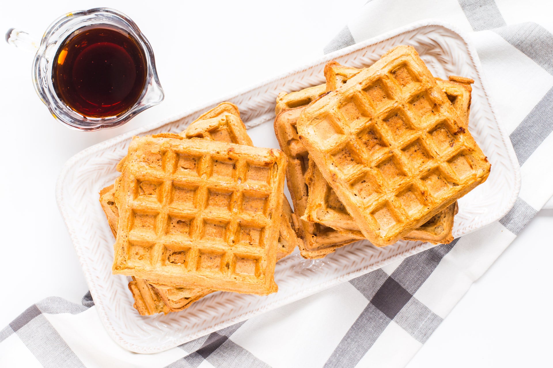 Vegan waffles sit stacked on a platter, with a dish towel underneath the platter. There is a small glass pitcher full of maple syrup be the waffles.