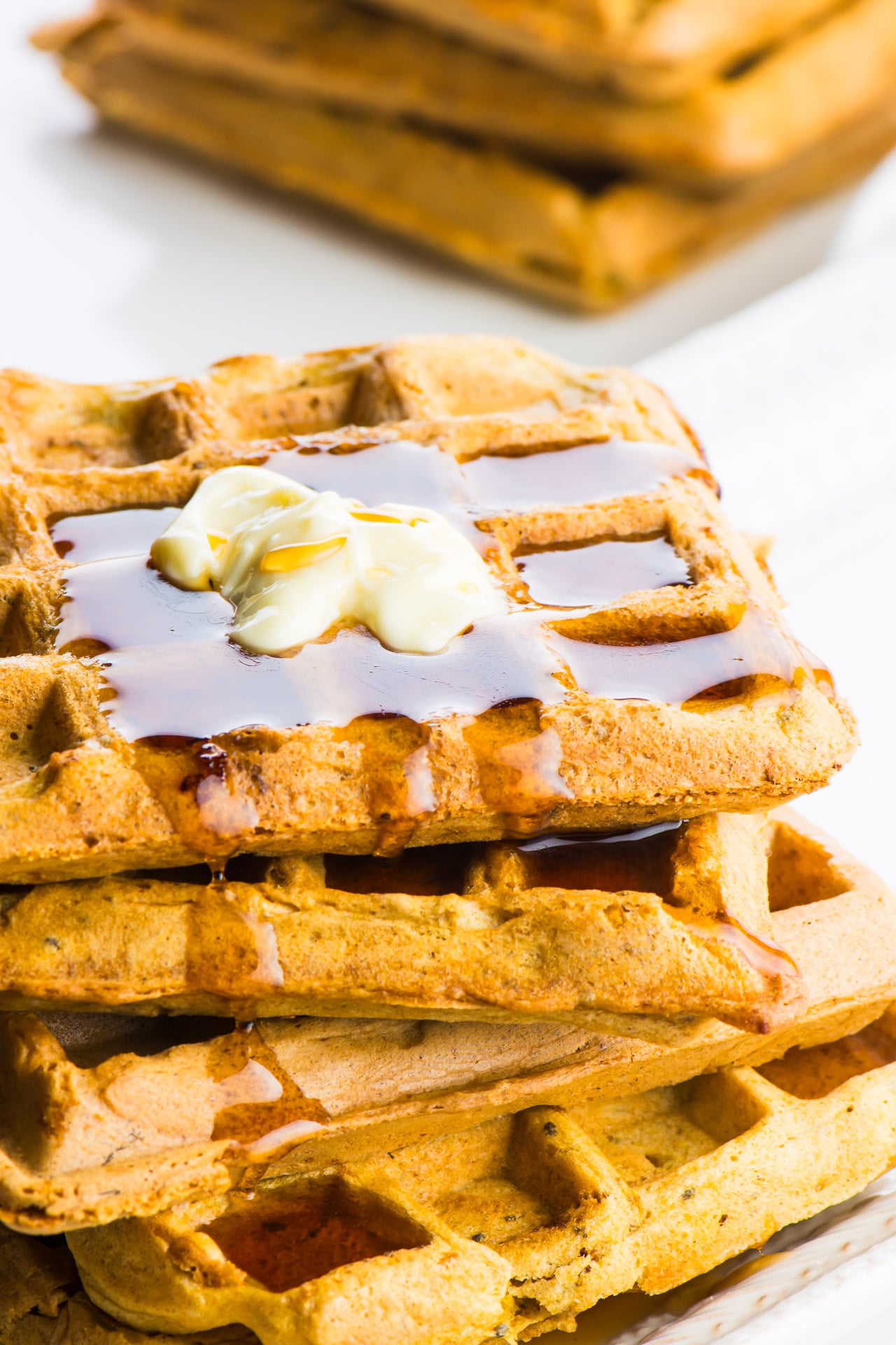 A stack of four whole wheat vegan waffles sit on a plate. There's melted vegan butter on the top waffle and it has maple syrup pooled in the waffle impressions on the top and oozing over the side. There are additional waffles in the background.