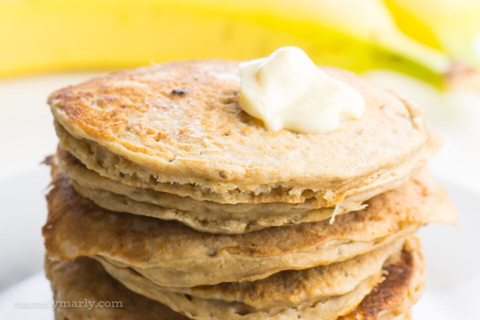 A close-up shot of banana pancakes with a pat of melted butter on the top. There are yellow bananas in the background.