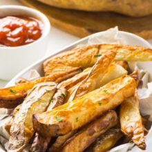 Some air fryer french fries are in a white bowl. A bowl of ketchup and raw potatoes on a cutting board are behind.