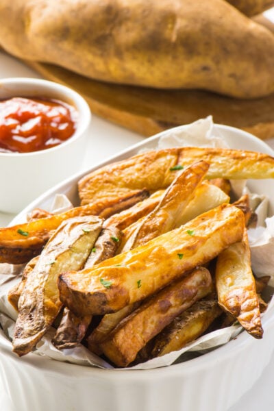 Some air fryer french fries are in a white bowl. A bowl of ketchup and raw potatoes on a cutting board are behind.