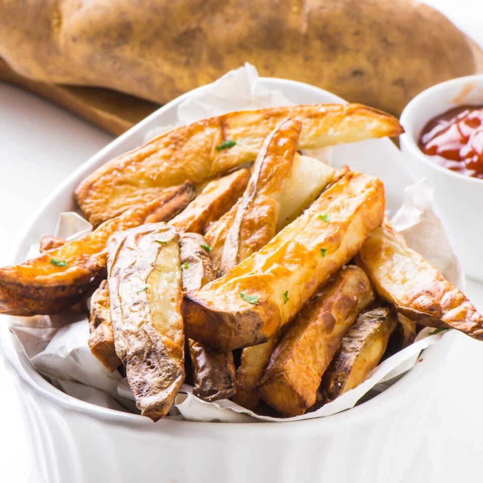 Air Fryer French fries are served up in a white bowl with ketchup and raw potatoes behind it.