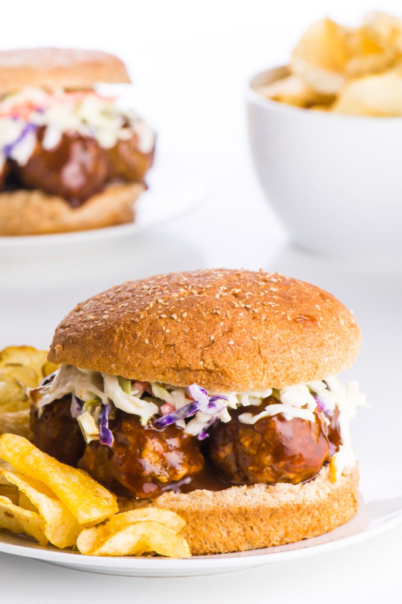 A lentil BBQ meatball sandwich with a whole wheat bun sits in the front with another sandwich behind it and a bowl of potato chips.