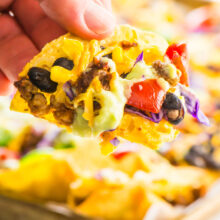 A hand reaches in with a baked vegan nacho taken from the pan behind it.