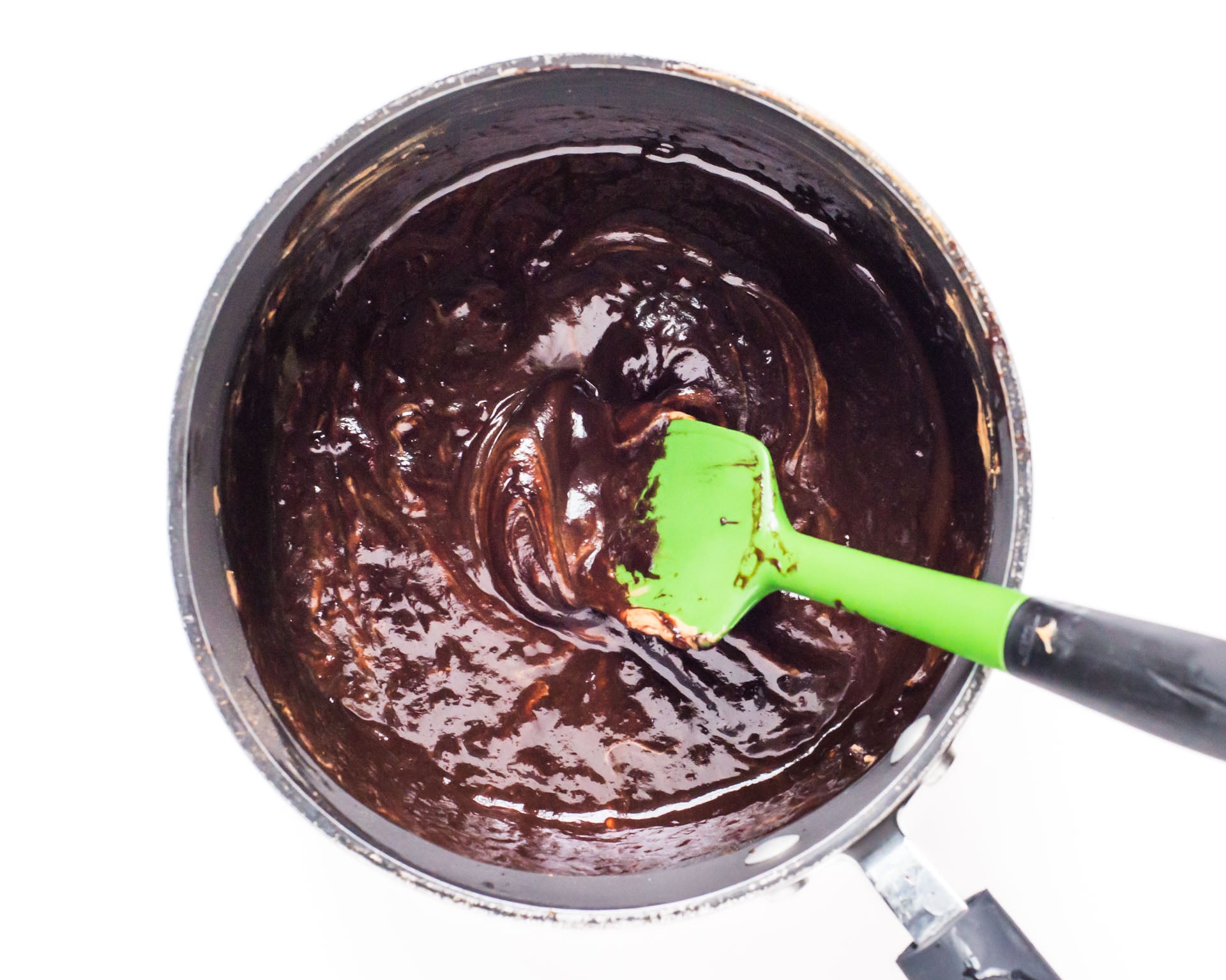 A saucepan is full of chocolate batter. A green spatula is stirring the ingredients.