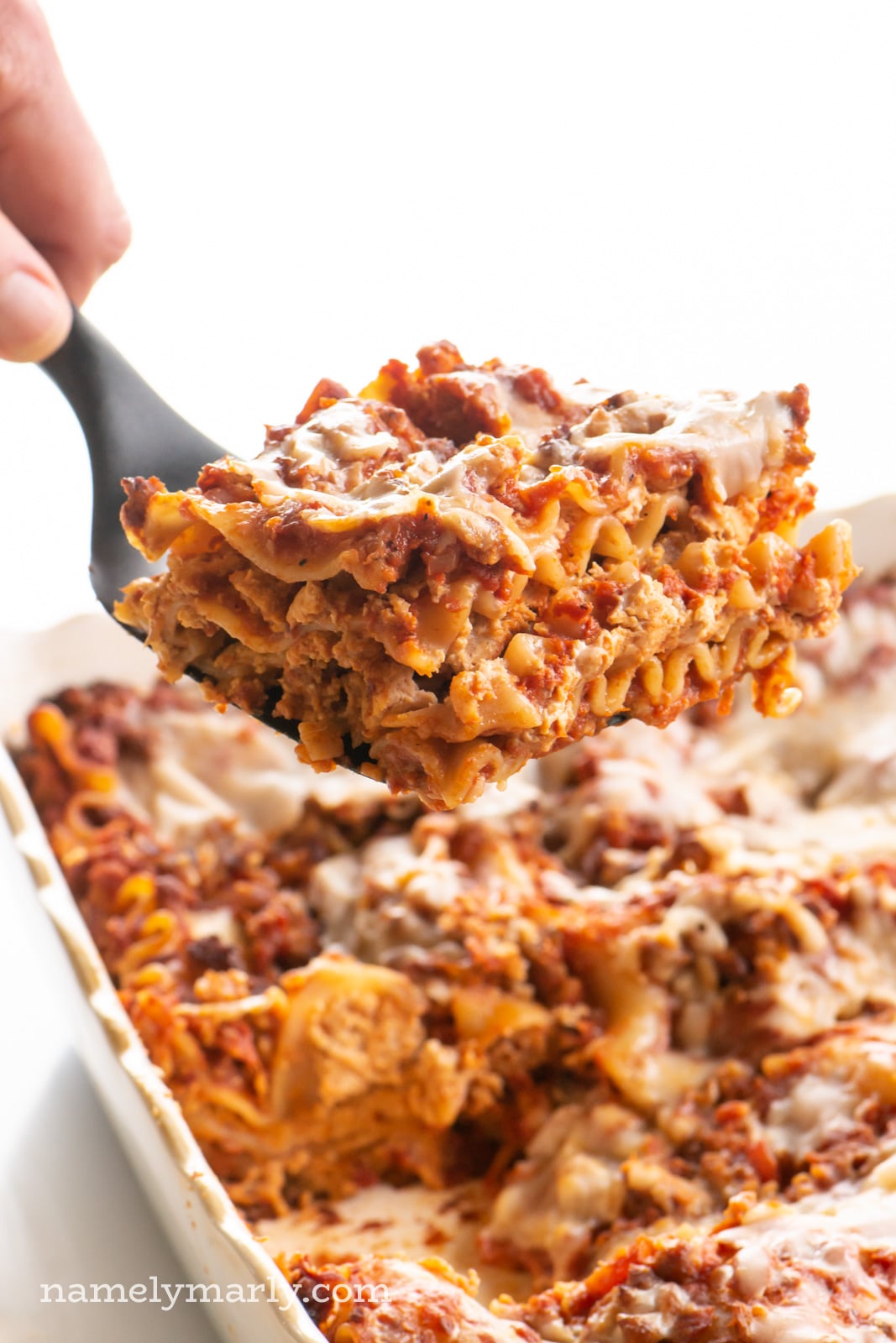 A hand holds a spatula with a slice of lasagna on it. The rest of the dish is behind it.