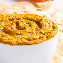 A bowl of carrot hummus is surrounded by chips with carrots in the background.