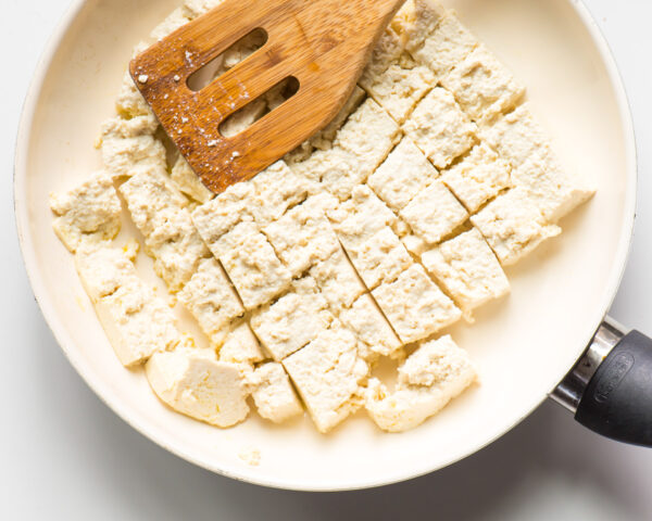 Tofu is in a skillet and it's broken into pieces by a wooden spatula.