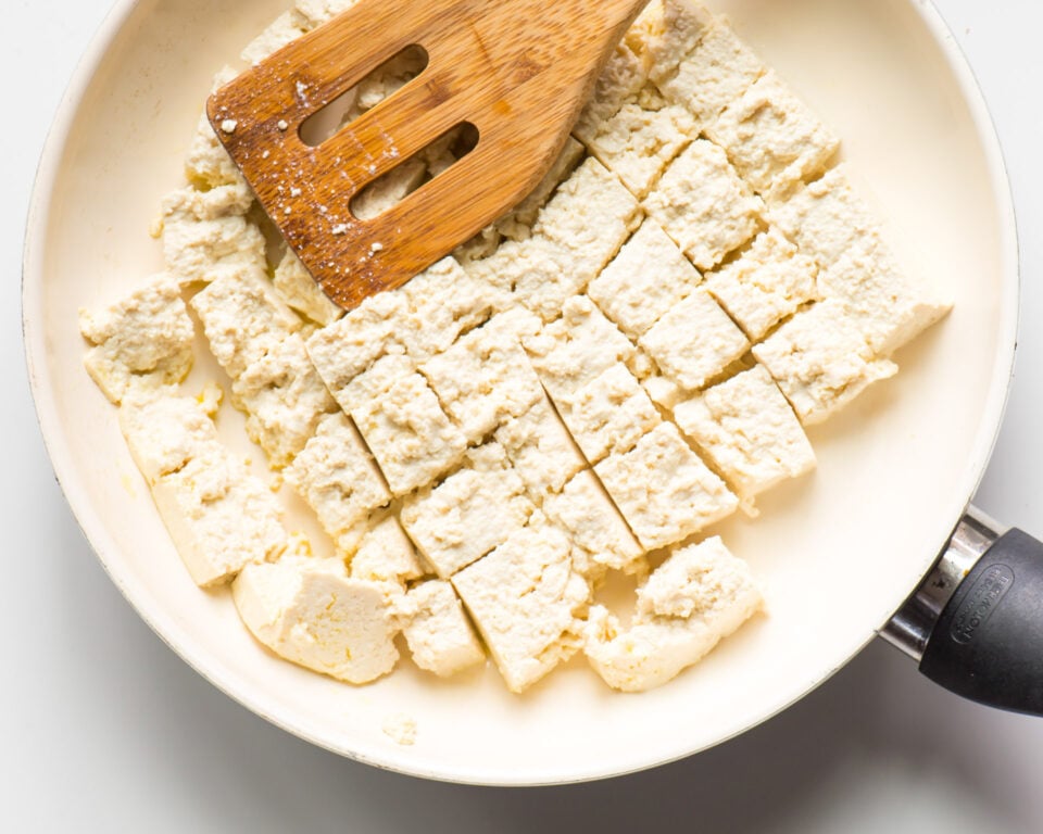 Tofu is in a skillet and it's broken into pieces by a wooden spatula.