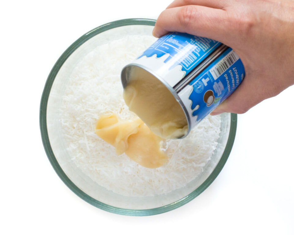 A hand pours coconut sweetened condensed milk into a bowl with shredded coconut.