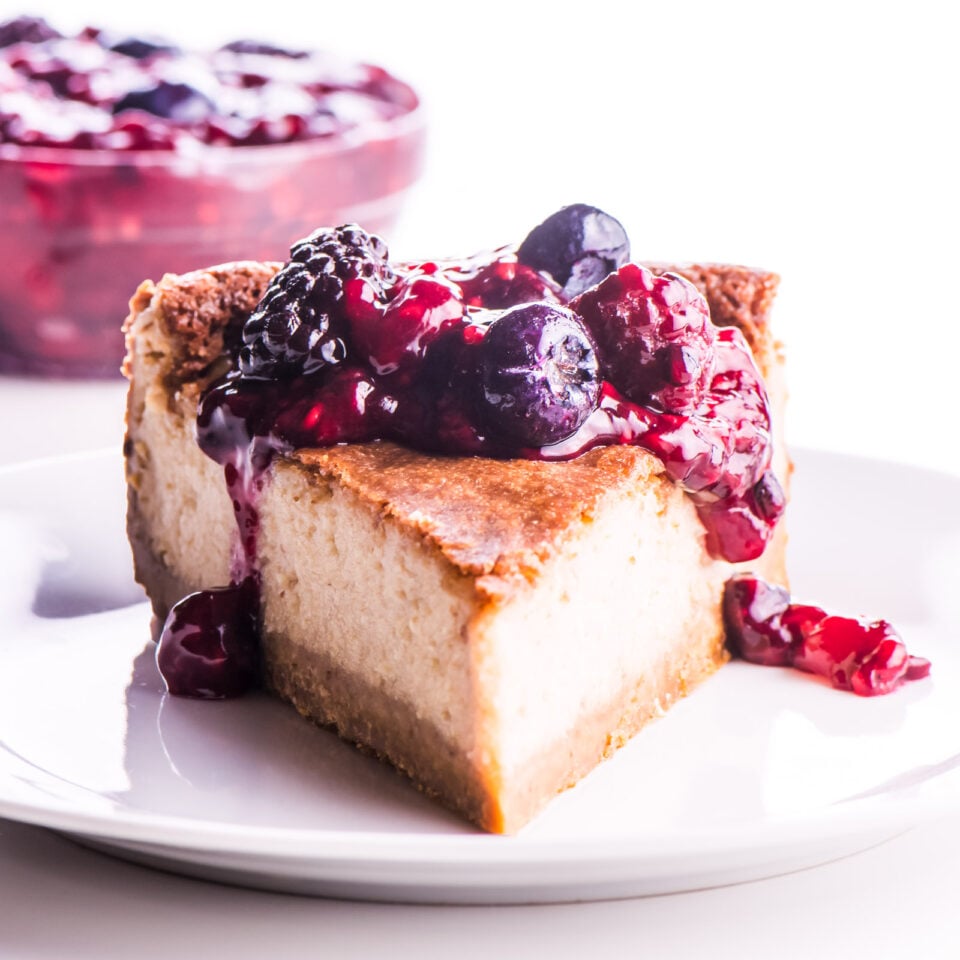 A slice of cheesecake with berry drizzle over the top and more berry topping in a bowl behind it.