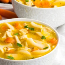 Two bowls of vegan chicken noodle soup sit one in front of the other with baby carrots beside the back bowl.