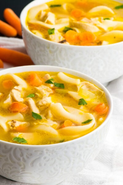 Two bowls of vegan chicken noodle soup sit one in front of the other with baby carrots beside the back bowl.