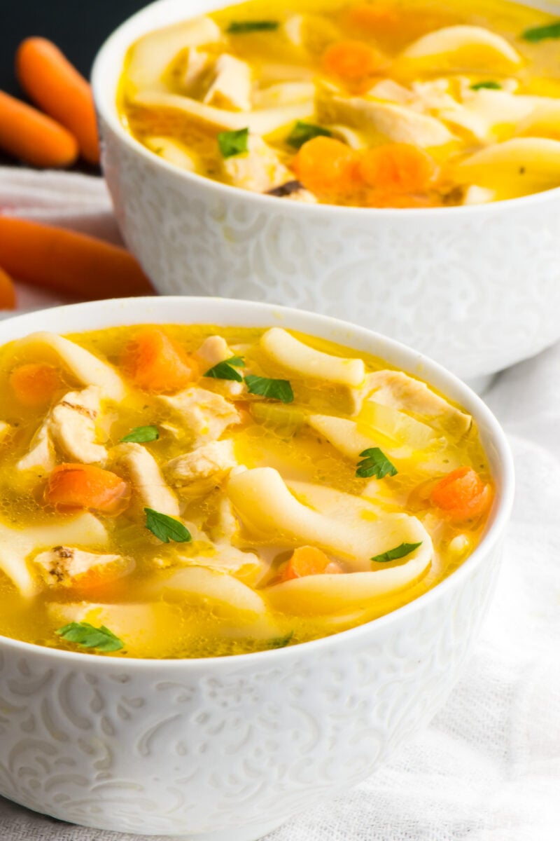 Two bowls of vegan chicken noodle soup sit one in front of the other with baby carrots beside the back bowl.