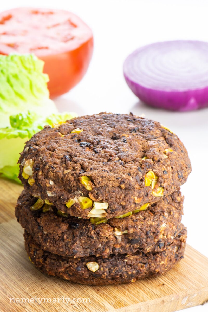 A stack of three sweet potato black bean burgers on a cutting board with lettuce, tomatoes, and onion behind it.