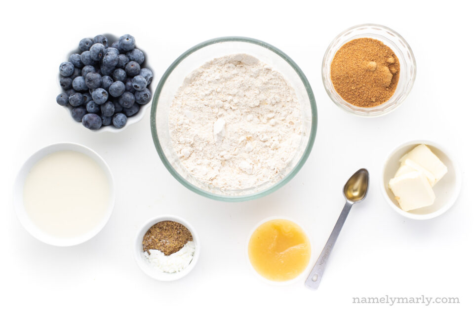 Looking down on bowls and measuring spoons full of ingredients like flour, coconut sugar, butter, blueberries, etc.