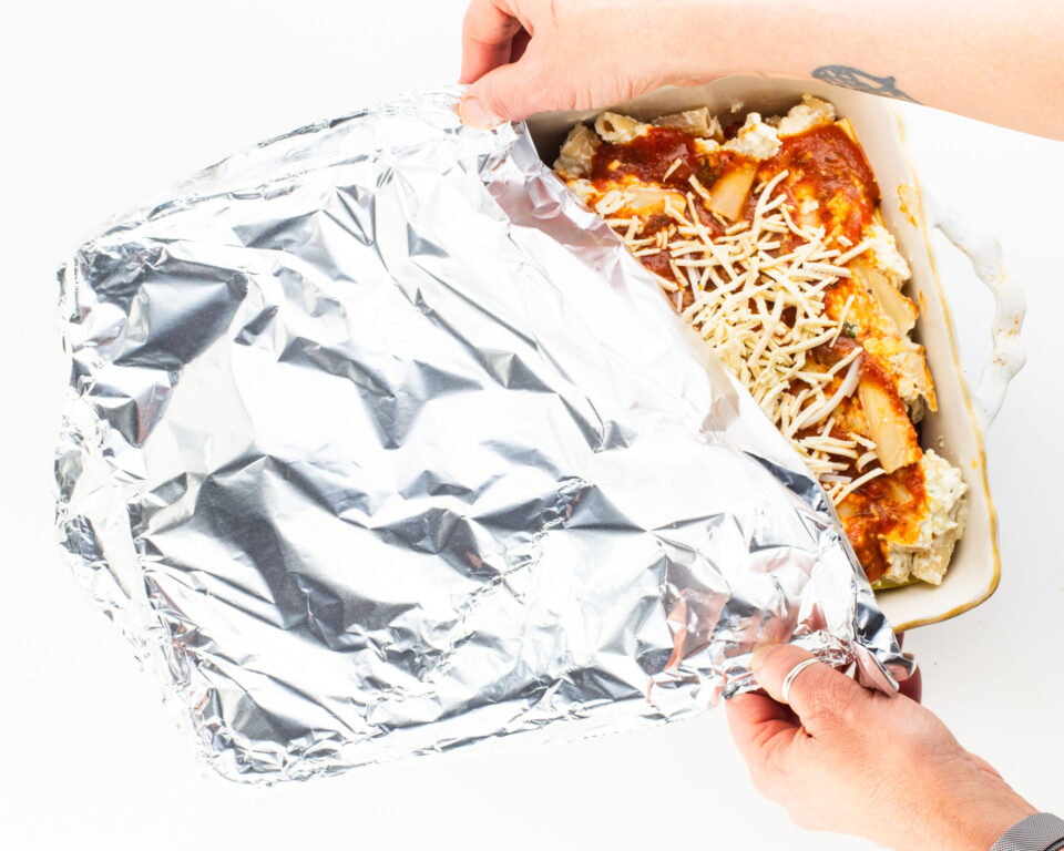Two hands reach in and cover an unbaked dish of meatless ziti with foil.
