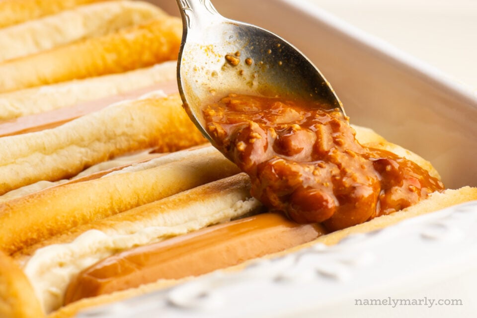 A spoon dishes out vegetarian chili over vegan hot dogs in a casserole dish.