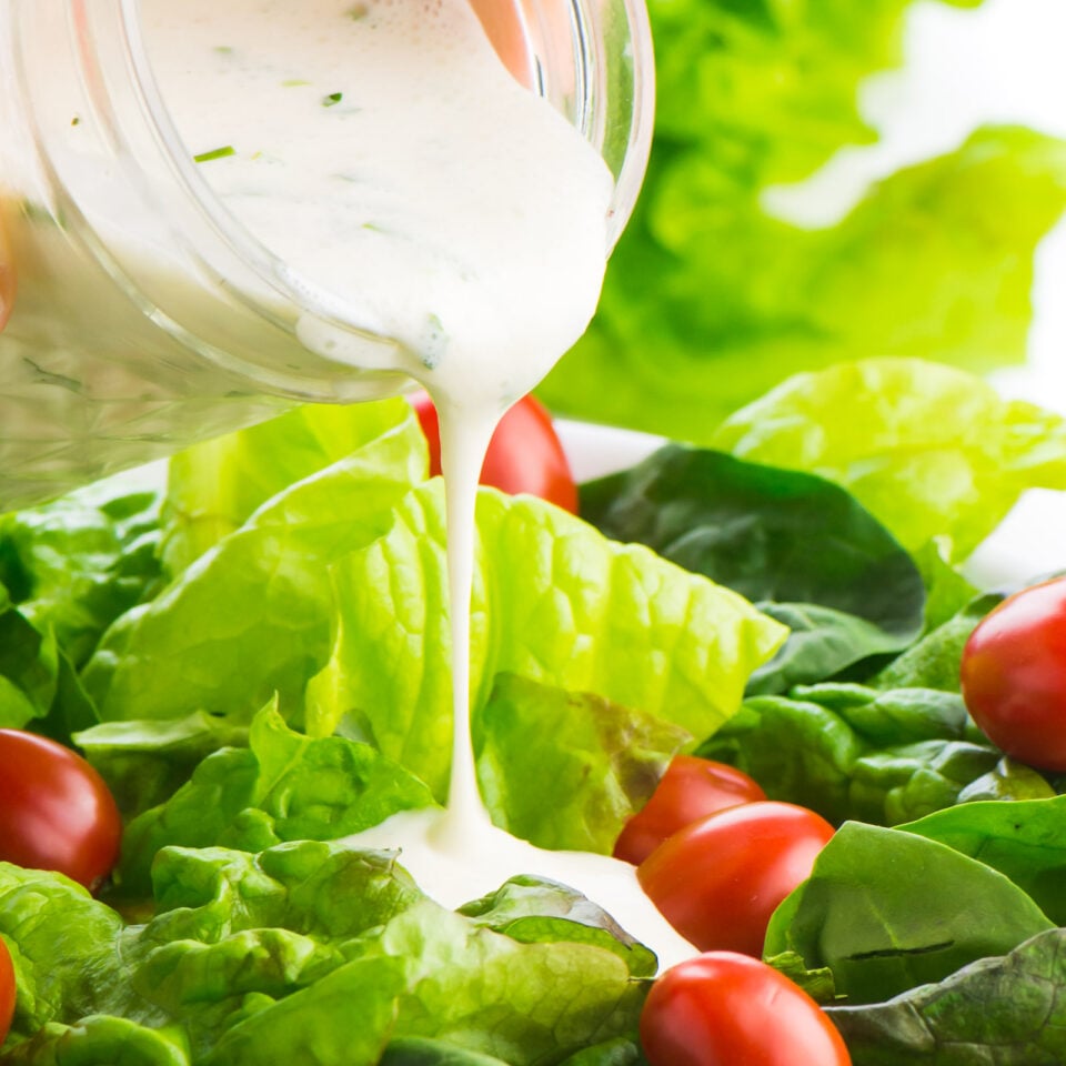 A mason jar full of vegan ranch dressing is pouring the dressing over a leafy, green salad with lots of bright red cherry tomatoes.