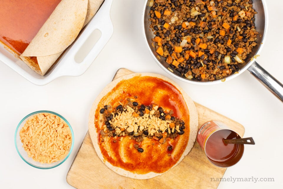 An unrolled enchilada sits on a cutting board with enchilada sauce and other ingredients nearby.
