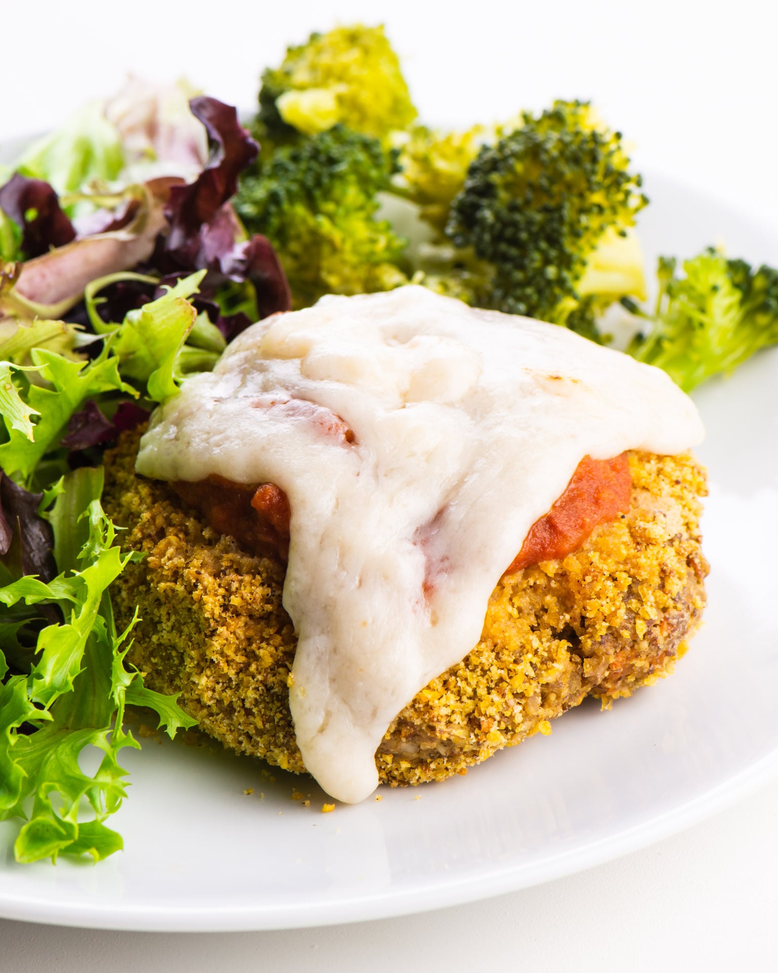 A vegan chicken parmesan patty on a plate with marinara sauce and melted vegan cheese. It sits beside a salad and steamed broccoli.