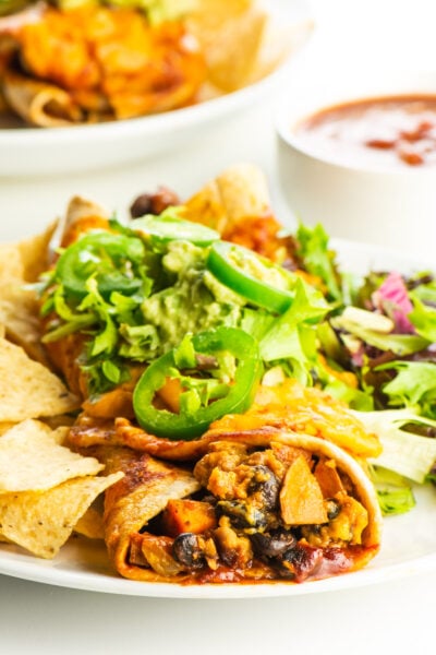 A vegan enchilada is topped with chopped lettuce and jalapeños with a bowl of salsa behind it.